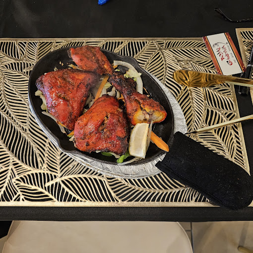 A sizzling platter of red tandoori chicken pieces, including drumsticks and thighs, garnished with sliced onions and carrots, and a lemon wedge, all resting on a black placemat with a gold leaf pattern.