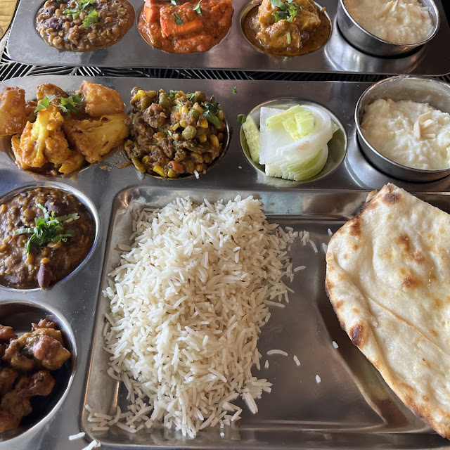 
A silver thali platter filled with a variety of Indian dishes, including rice, naan bread, several small bowls of different curries, a vegetable dish, and a bowl of raita, all neatly arranged.