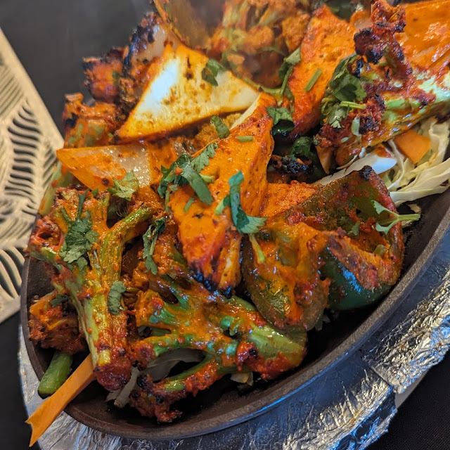 A sizzling platter filled with an assortment of tandoori-marinated vegetables, including broccoli, cauliflower, bell peppers, and paneer, garnished with fresh cilantro and steam rising from the hot dish.
