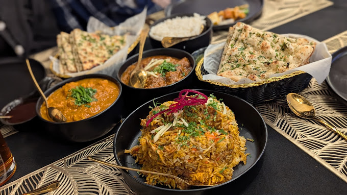 A vibrant spread of Indian dishes on a black tablecloth with a gold patterned placemat, featuring a large bowl of biryani with red onion and herbs, two bowls of curry, baskets of naan bread, and a bowl of rice.