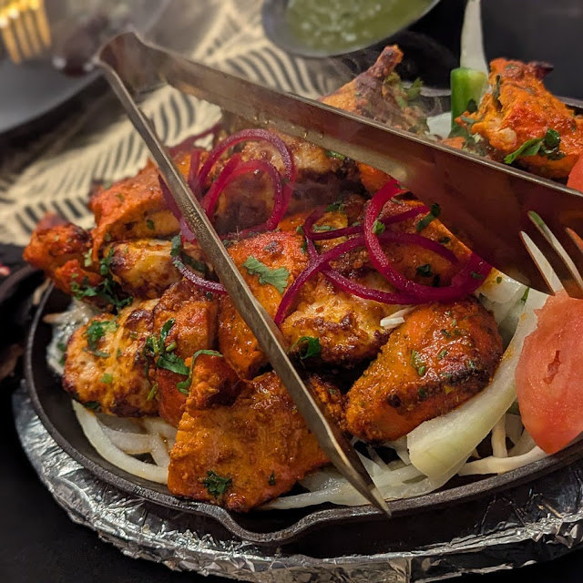 A sizzling cast iron platter filled with assorted tandoori chicken pieces, garnished with red onion slices and cilantro, served alongside watermelon slices, a lime wedge, and a small bowl of green chutney, with tongs resting on the chicken.
