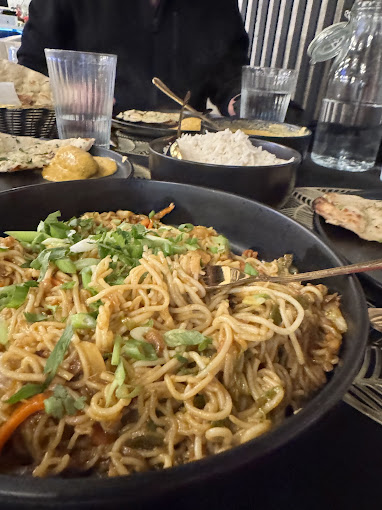 
A close-up of a black bowl filled with noodles, possibly chowmein, topped with chopped green onions and other vegetables, with a fork resting in the dish. In the background, other Indian dishes like naan and rice are visible on a dining table.